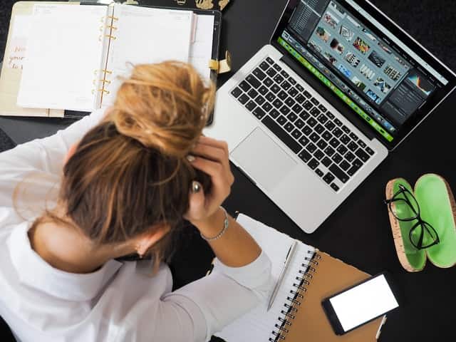 A woman at work feeling stressed looking down and holding her head in front of her laptop and notebook
