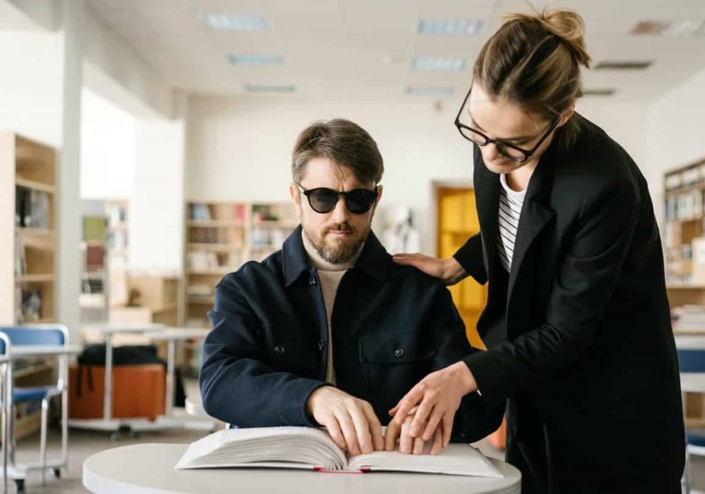 Blind man reading brail on desk with a woman helping him