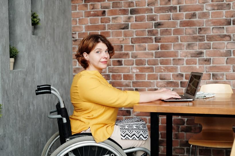 A wheelchair user is sitting at a table and working on her laptop